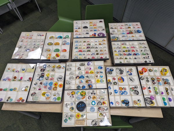 A photograph of 12 archival trays containing hundreds of badges in lots of different colours. You cannot make out the detail of any of the badges from the photograph 