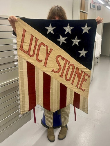 A person holding a suffrage banner while in LSE Library Archives. The banner is similar to the stars and stripes flag of the USA and has 'Lucy Stone' written on it. 