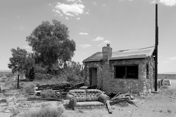 A small (one room?) stone house, damaged by fire, with a couch sitting in front of it. A tree sits behind and to the left of the house. Brush and the barren surrounding landscape suggest the desert.