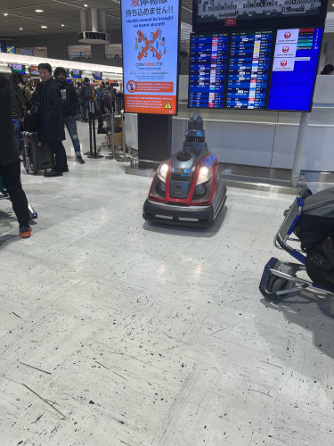 A small robotic cart (purpose not clear!) sits in a relatively clear area of the airport hall, as people have tried to meet its demands to scat. 