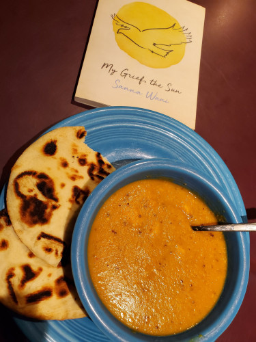 Poetry collection My Grief, the Sun by Sanna Wani (House of Anansi Press), with the hand drawn image of a bird flying across a bright sun, sits next to a bowl of bright mulligatawny soup in a blue bowl, accompanied by toasted naan