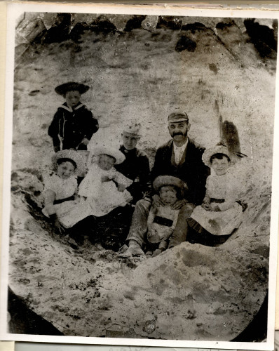 A photograph of George Lansbury 
in a flat cap surrounded by his young children in what looks like a sandpit on the beach