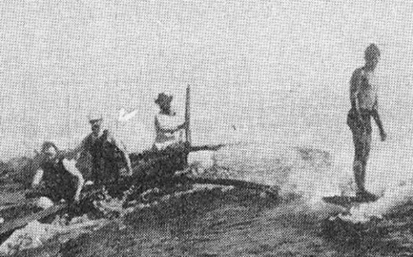 Very grainy image of Jack London surfing, Waikiki, 1907, next to three men in an outrigger.