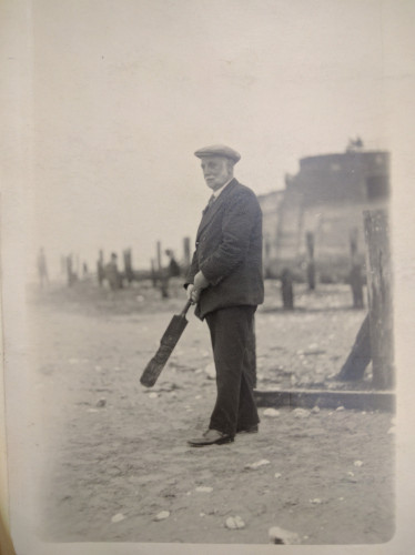 A black and white photograph of George Lansbury in a suit and hat, holding a cricket bat as if about to hit a ball. It looks like he is standing on a beach somewhere. 