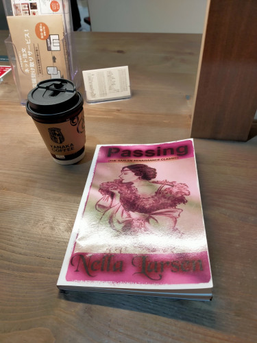 The photo is of a coffee counter with a pink book that has an illustration of a flapper style with a massive feather boa & a headband with short hair looks off to the left. Since the novel is about "passing" for white, the illustration is intended to be of a light-skinned Black woman. To the left is a Yanaka Coffee logo-ed sleeved paper coffee cup with a black lid