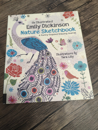 A cream colored book with birds and flowers across the front. One bird  is a large peacock. Book is titled The Illustrated Emily Dickinson Nature Sketchbook: A Poetry Inspired Drawing Journal. Sitting on a wood table.