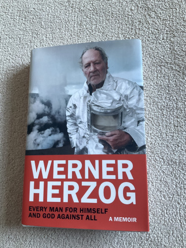 Cover of Every Man for Himself and God Against All - Werner Herzog. Cover photo depicts Herzog, a white man in his 70s, wearing a silver protective suit, holding the headpiece in his hands. I think he is in a volcano crater.