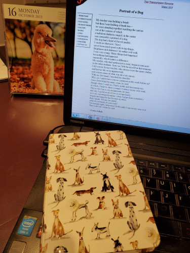  The poem "Portrait of a Dog" by Louise Glück appears on a computer screen on the web site of The Threepenny Review, with a notebook with cartoon dogs on the cover sitting on the computer keyboard. A dog calendar is in the background.