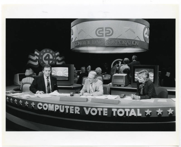 Local Minneapolis TV set on election. night. Black and white photo with two men and one woman on the panel behind a desk that says "Computer Vote Total." Dark background with computers and workers
 in background on the news set.