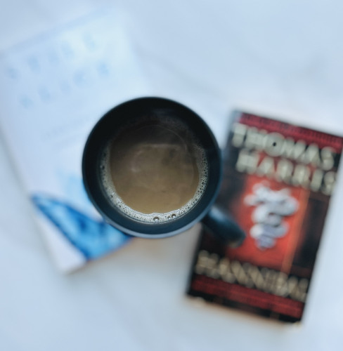 An in-focus black cup of coffee with a smidge of steam coming off of it, sitting a top of two books (Still Alice by Lisa Genova and Hannibal by Thomas Harris, which are purposefully out of focus).