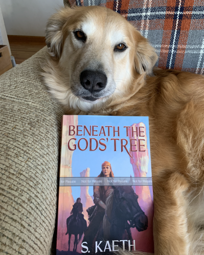 A golden collie mix propping up a novel, Beneath the Gods' Tree by S. Kaeth, which shows a woman riding a horse through a rocky desert.