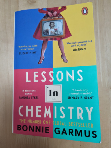 Front cover of the paperback edition of "Lessons in Chemistry" by Bonnie Garmus. The background is divided into four equal, colourful sections and there is the bottom half of a female figure at the top of the cover. She is wearing a fitted dress or coat and heels and carrying an old-fashioned looking TV which is showing an image of a woman in the middle of a chemistry experiment. 