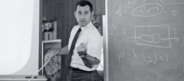 Black and white image of Prof Kleinrock giving a lecture in a classroom flanked by an overhead projector and a chalkboard. 