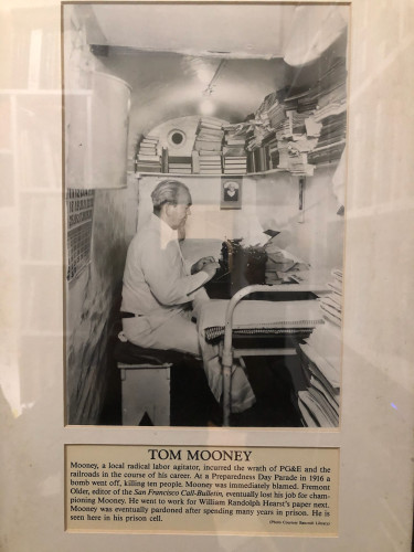 Photography of a graying, balding Tom Mooney in his tiny prison cell, typing on a typewriter that is positioned on his bed. He is surrounded by stacks of books and papers.