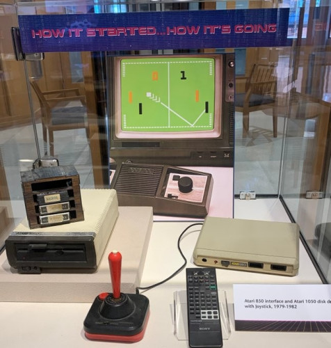 Color image of display case with Atari game system from the late 70s & early 80s including screen, dial controllers, cartridges of games, joysticks, a disk drive....