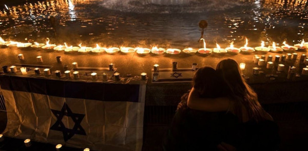 People embrace next to an Israeli flag and a large circle of candles around a circular fountain or pool of water at nighttime.