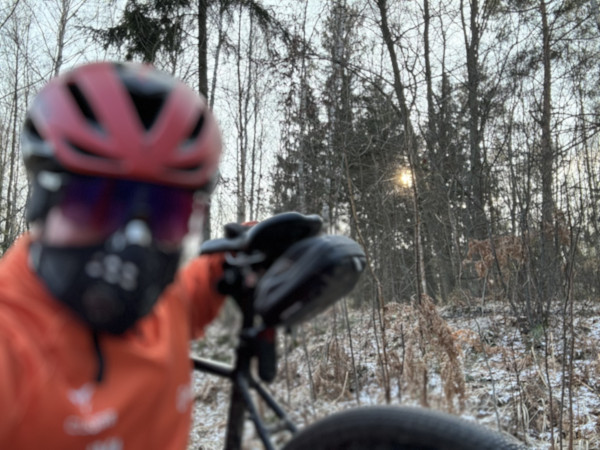Rowerzysta w kasku i częściowo zakrywający twarz robi sobie selfie na rozmytym tle zaśnieżonego lasu i słabego słońca prześwitującego przez drzewa.