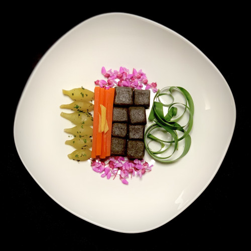 Black plate, white roundish square plate turned so the ends pointing to each side of the pic. Vegetables are arranged in an organized colorful cluster on the plate.  On the left are steamed dashi-infused daylily roots that look like dark yellow bowling pins and are pointing to the left of the plate. Flecks of culantro is sprinkled on top. Next are steamed dashi-infused thin slices of carrots with thin slices of ginger laying on top. Next are near black noodle squares of live oak acorn noodles with black sesame seeds and to the right are daylily leaves curled into ribbons. Above and below are rosebud flowers. 