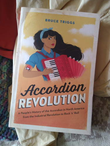 "Accordion Revolution: A People's History of the Accordion in North America from the Industrial Revolution to Rock and Roll", a book by Bruce Triggs. It's sitting on a chair. The cover features an illustration of a long-haired, brown-skinned woman wearing a blue button-up shirt with sleeves rolled up, revealing an accordion & heart tattoo on her bicep, and playing a red piano accordion.