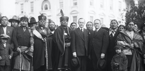 Men and women in a mix of suits and traditional dress stand in front of the White House. 