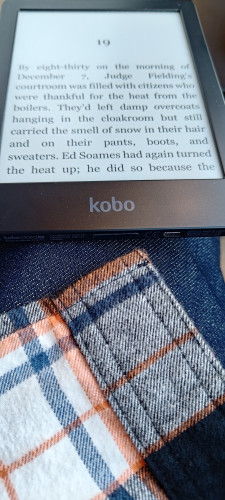 A Kobo e-reader resting my leg. Blue jeans and the corner of a flannel shirt are visible. 