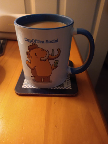 Blue cupofteasocial mug on a wooden bedside cabinet
The mug is cream coloured with a blue handle on the right inside of the mug is blue, at the top of the mug the words cupofteasocial above a picture of Tusky 