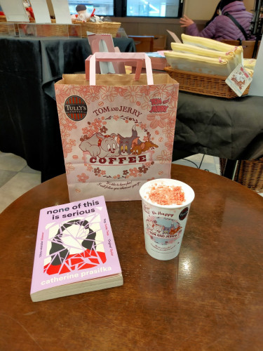 The photo is of a round reddish brown table. On the table is the paperback book mentioned in the toot. It has a purple frame of a shattered illustration of a faceless woman with black hair and a red top, the cracks being in purple too. To the right is a paper latte cup with strawberry chocolate flakes on top of whipped cream. The white paper cup says BE HAPPY above a circular illustration of TOM AND JERRY characters, a light grey dog, dark grey cat, & red mouse. Behind is a pink paper bag with the same illustration. The characters are looking at the word COFFEE below them. The bag has pink flowers all over it and the orange/green vertical striped TULLY'S logo is in the left hand corner. You can see the back of a display case beyond the table and blurry patrons sitting to the left.