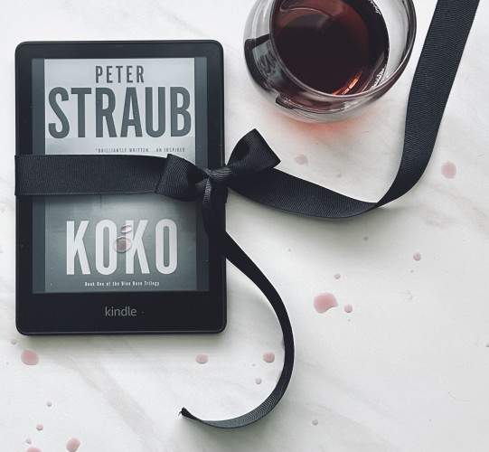 My Kindle, featuring Koko by Peter Straub, has a ribbon tied around it. A small glass of red wine is toward the top of the pic with drops splattered. 