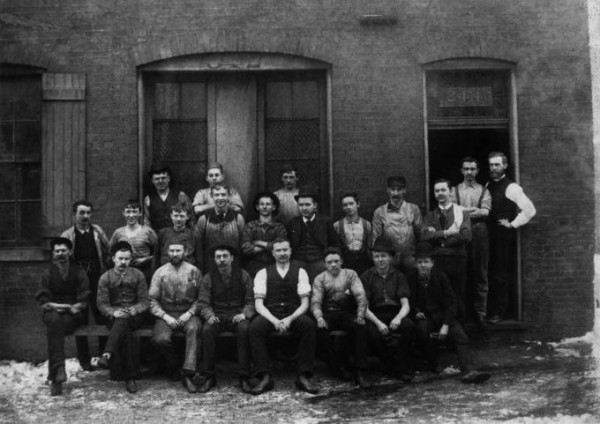 Image of about two dozen workers in front of their factory building with the first row seated and the back two rows standing.