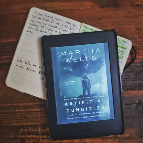 An ebook reader, on top of a pocket notebook, in top of a wooden table top. The cover of Martha Well's book Artificial Condition is on the eReader.