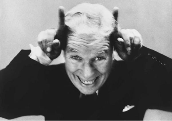 An elderly Charles Chaplin, grinning broadly, faces the camera, with his index fingers pointing up on either side of his head, making him seem like a devil.
