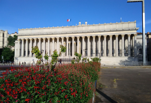 Au premier plan, un parterre de fleurs rouges et d'arbustes verts, avec le palais de justice en perspective.
La façade est néo-classique avec, sur toute sa largeur, des escaliers, 24 colonnes surmontées de chapiteaux corinthiens, un fronton plat et décoré.
Au-dessus de l'édifice, un drapeau français.
A l'arrière, le sommet de la basilique de Fourvière. A gauche et à droite, d'autres bâtiments. Le ciel est bleu et lumineux.
