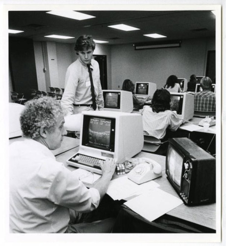 B&W image of a man sitting at a computer and TV in the foreground as another man stands next to his desk looking over election returns. 