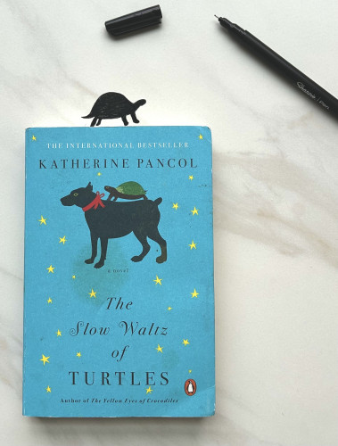 A book with a dog and turtle on the cover, titled, The Slow Waltz of Turtles, by Katherine Pancol. I have drawn a little turtle across the top of where the book is laying. My ink pen and cap are off to the right. 