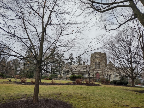 Photo: Bare trees in the foreground in front of a single story Gothic building with a small tower above an arched red front door. There is a winter barren lawn stretching from the trees to the building. Off to the left of the building in the background are several tall pine trees. The sky is light gray as a result of cloud cover.