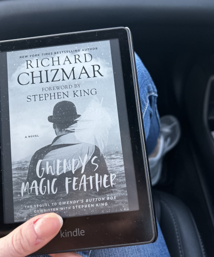 Me, holding my Kindle, which has Gwendy’s Magic Feather by King and Chizmar on it. My leg and shoe are in the background 