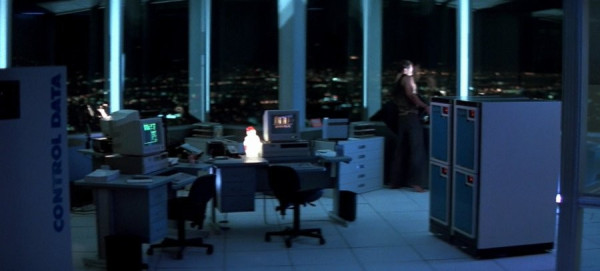 Screen shot from the 1988 film Die Hard bluish tone night image of high-rise open office room with floor to ceiling windows looking out on a city scape a Control Data Cyber 180 on the left with desks and workstation monitors in the middle.