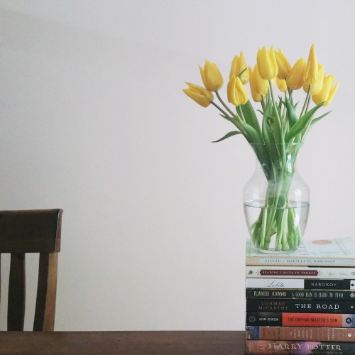 My table, with a small stack of books and a vase of yellow tulips is on top of the books. A chair peeks out on the left.