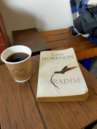 The photo is of a brown faux wood table on which is the worn white paperback book with a bright green yellow black & long skinny orange beak bird of paradise. To the right is a paper cup of black coffee. The cup is white interior with brown exterior with the coffee shop's logo of a 'phone' made from 2 paper cups & string